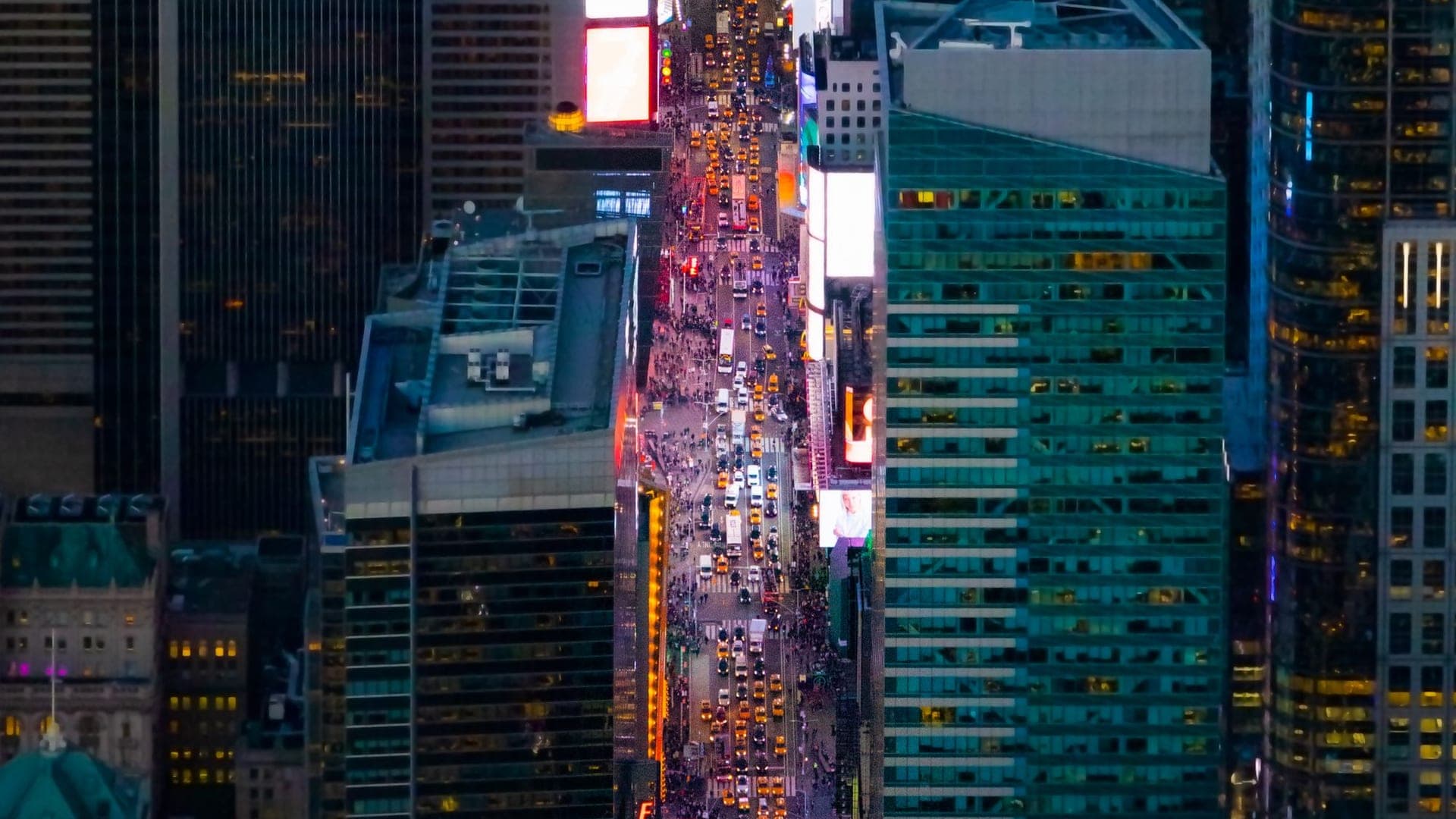 An aerial night view down a canyon of glowing towers and streaming traffic — a city running as one live grid of light and data.